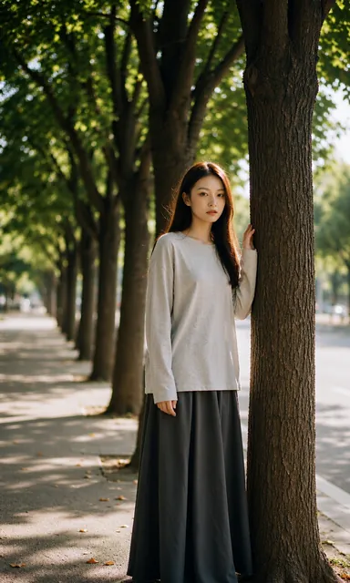 Woman in a long dark skirt standing in the shade of a line of trees with one hand against a trunk