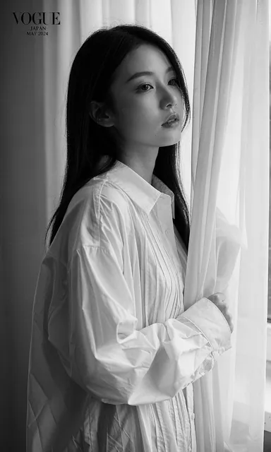 Black and white side profile portrait of a woman in a white cotton shirt beside a tall window with strong directional light